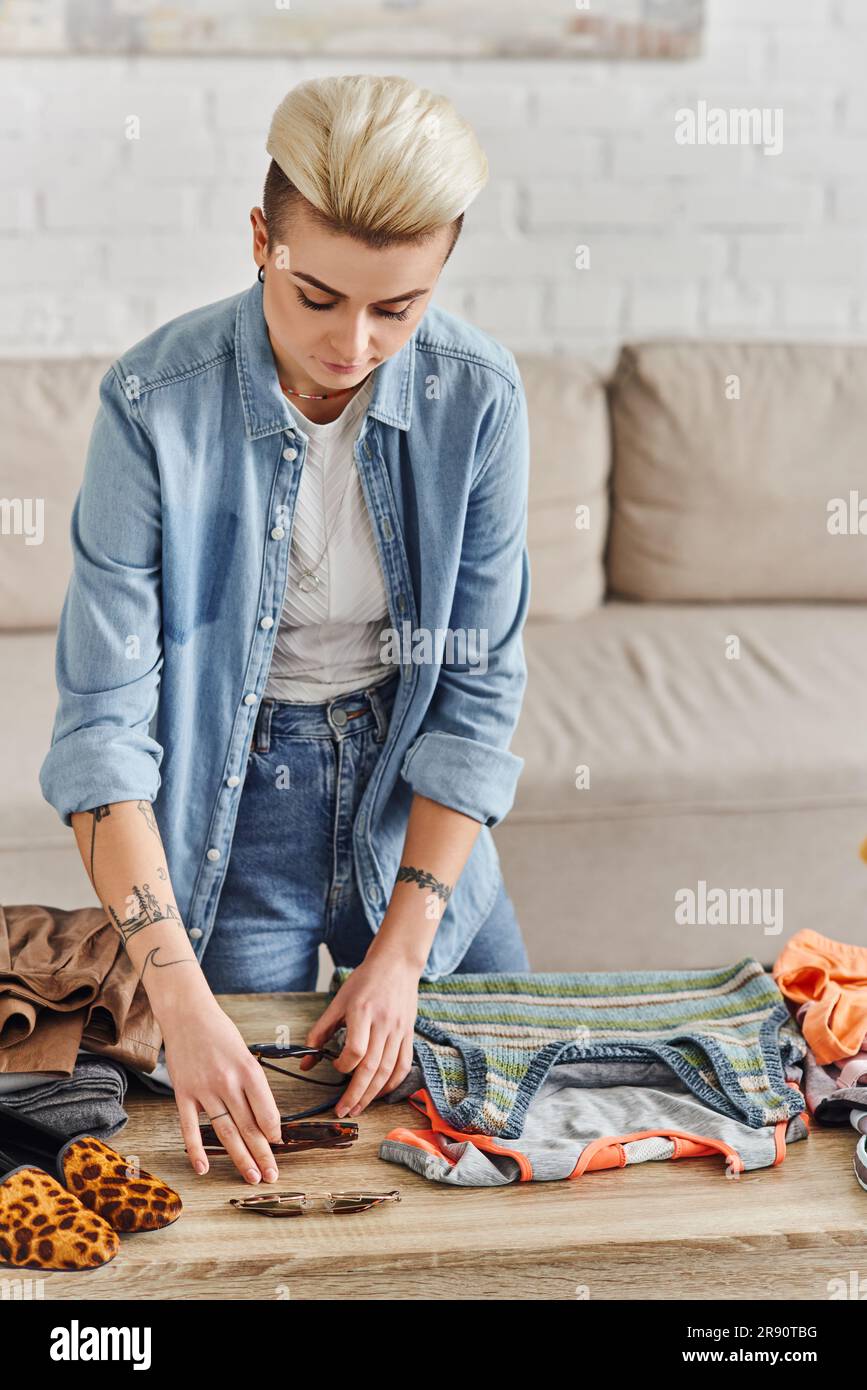 donna alla moda e tatuata che mette gli occhiali da sole sul tavolo mentre smistano oggetti e vestiti per il prediletto a casa, economia circolare, vita sostenibile Foto Stock