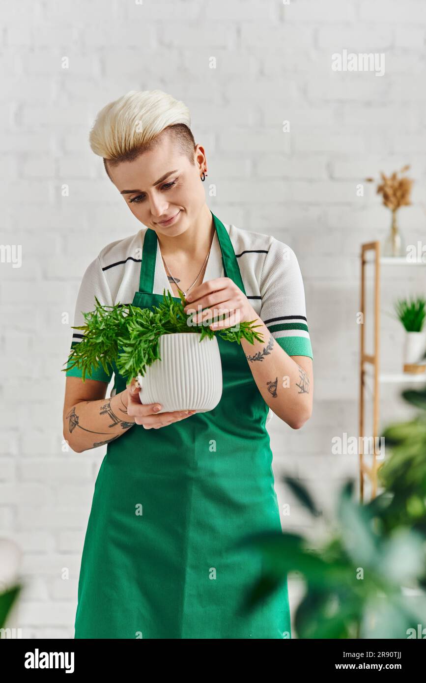 cura delle piante, giardinaggio interno, donna tatuata sorridente in grembiule verde che tocca piante in vaso naturali mentre si trova in un soggiorno moderno, sostenibile Foto Stock