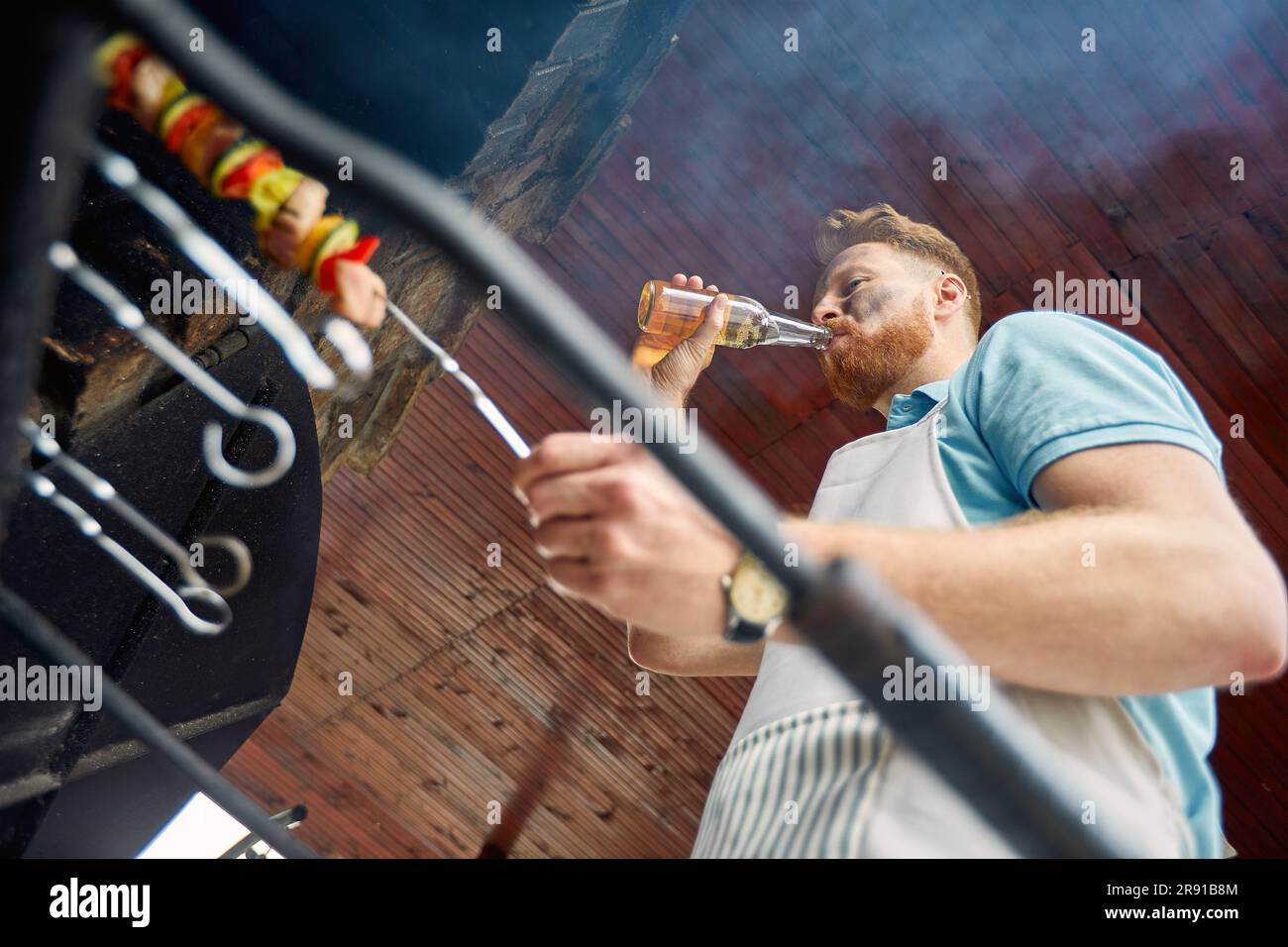 Lo chef si prende cura della griglia con sicurezza. Con una mano che gira sapientemente carne e verdure, l'altra contiene una bottiglia di birra fredda Foto Stock