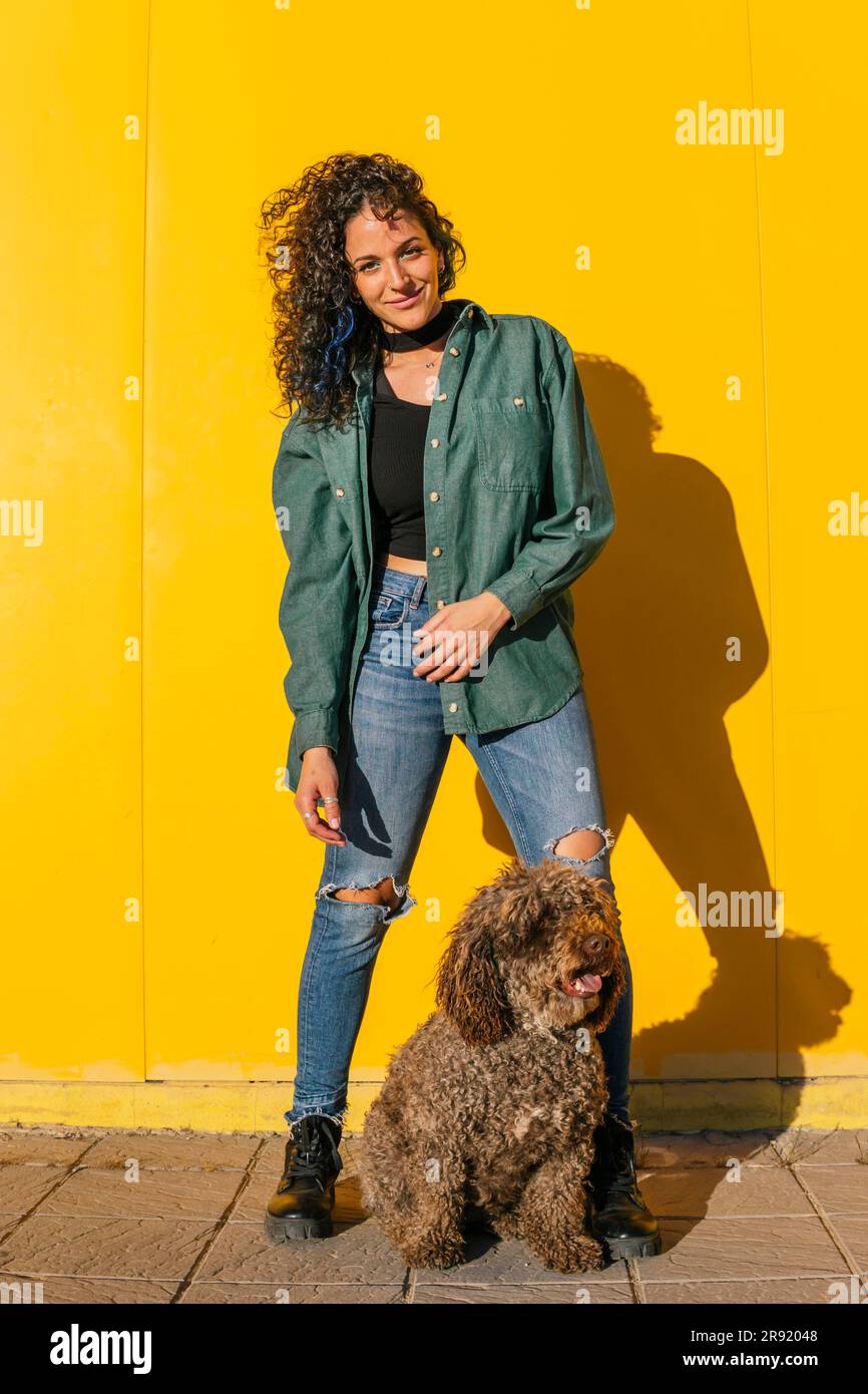 Donna sorridente in piedi con cane d'acqua di fronte al muro giallo Foto Stock