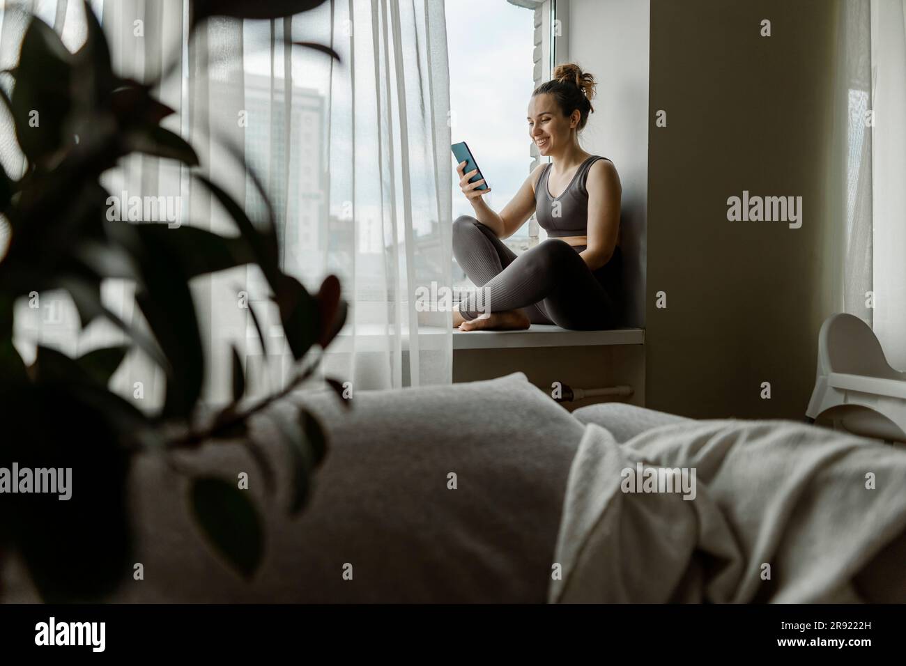 Una donna che invia messaggi di testo con il telefono seduto sul davanzale a casa Foto Stock