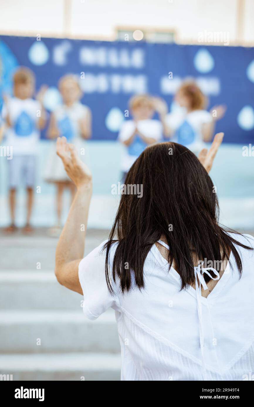 Schiena di una donna in primo piano battendo le mani guardando un gruppo di bambini sul palco all'aperto su sfondo sfocato Foto Stock