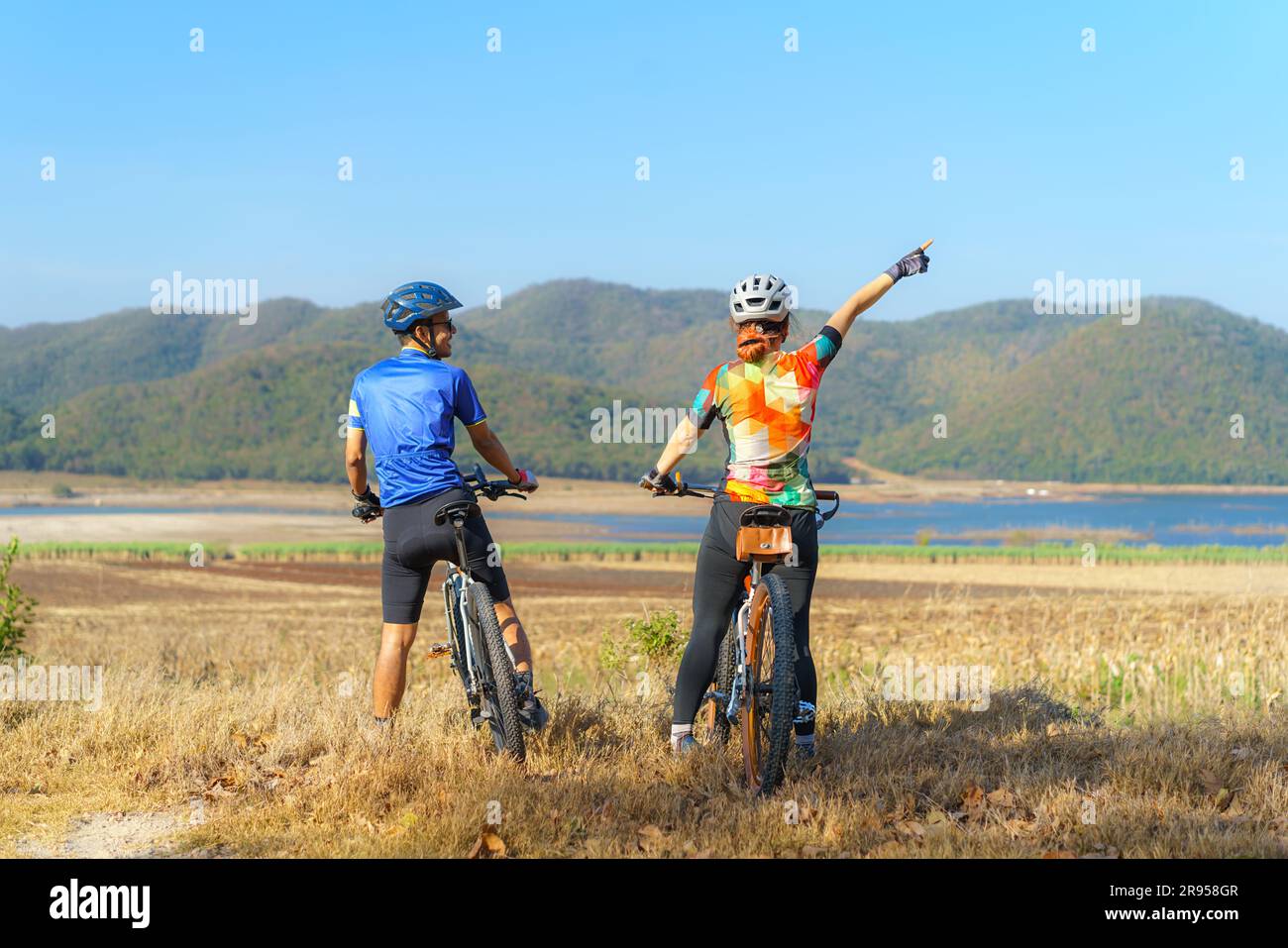 Coppia di ciclisti asiatici che guardano il lago e la montagna e chiacchierano mentre si prende una pausa dal giro in bicicletta mattutino Foto Stock