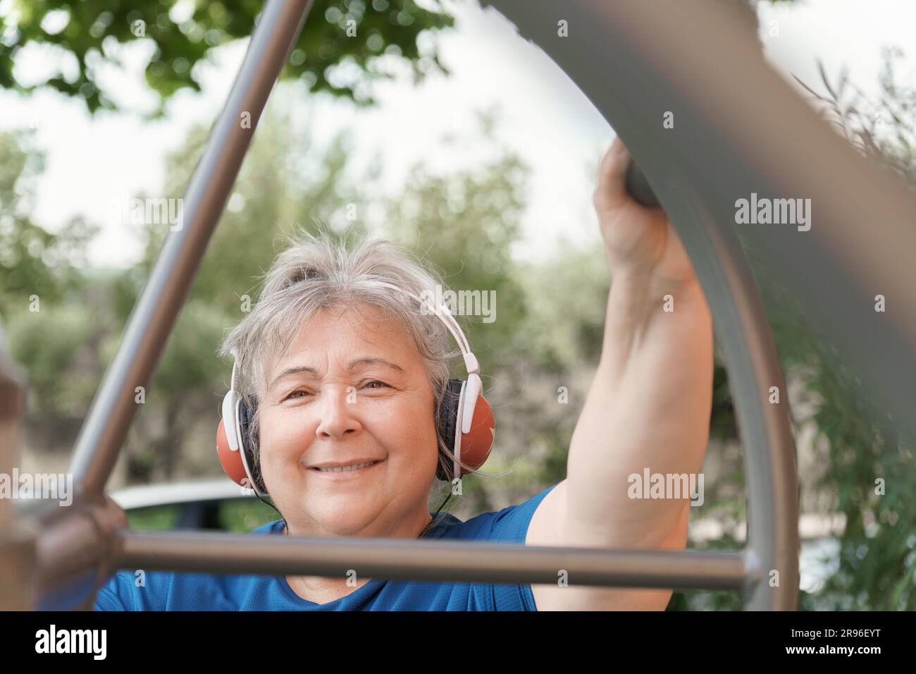 Donna anziana obesa attiva in abbigliamento sportivo che si allena su una macchina per l'esercizio del parco mentre ascolta la musica sulle cuffie Foto Stock