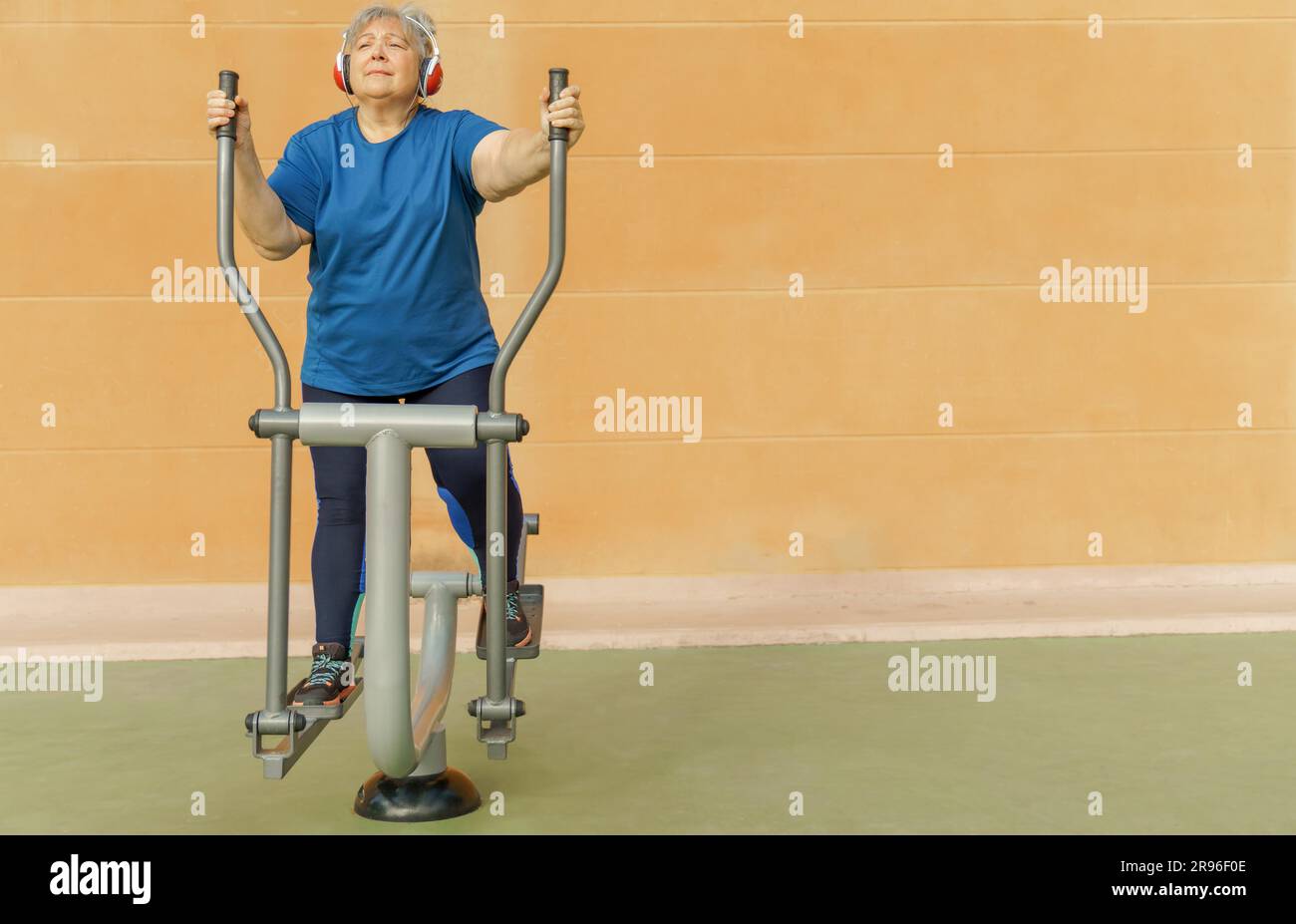 Donna anziana obesa attiva in abbigliamento sportivo che si allena su una macchina da parcheggio mentre ascolta la musica sulle cuffie Foto Stock