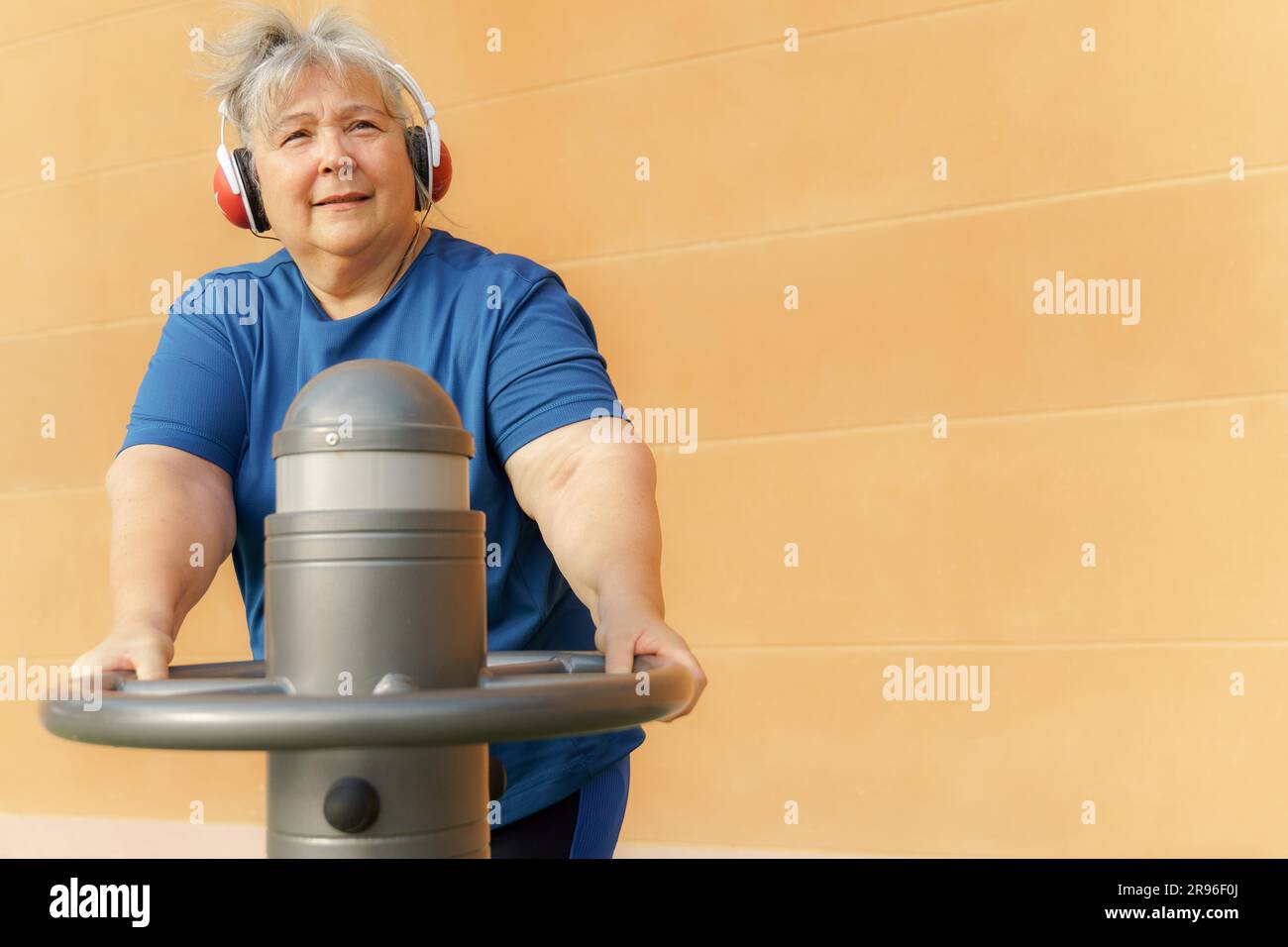 Donna anziana obesa attiva in abbigliamento sportivo che si allena su una macchina da palestra in una palestra del parco mentre ascolta la musica con le cuffie Foto Stock