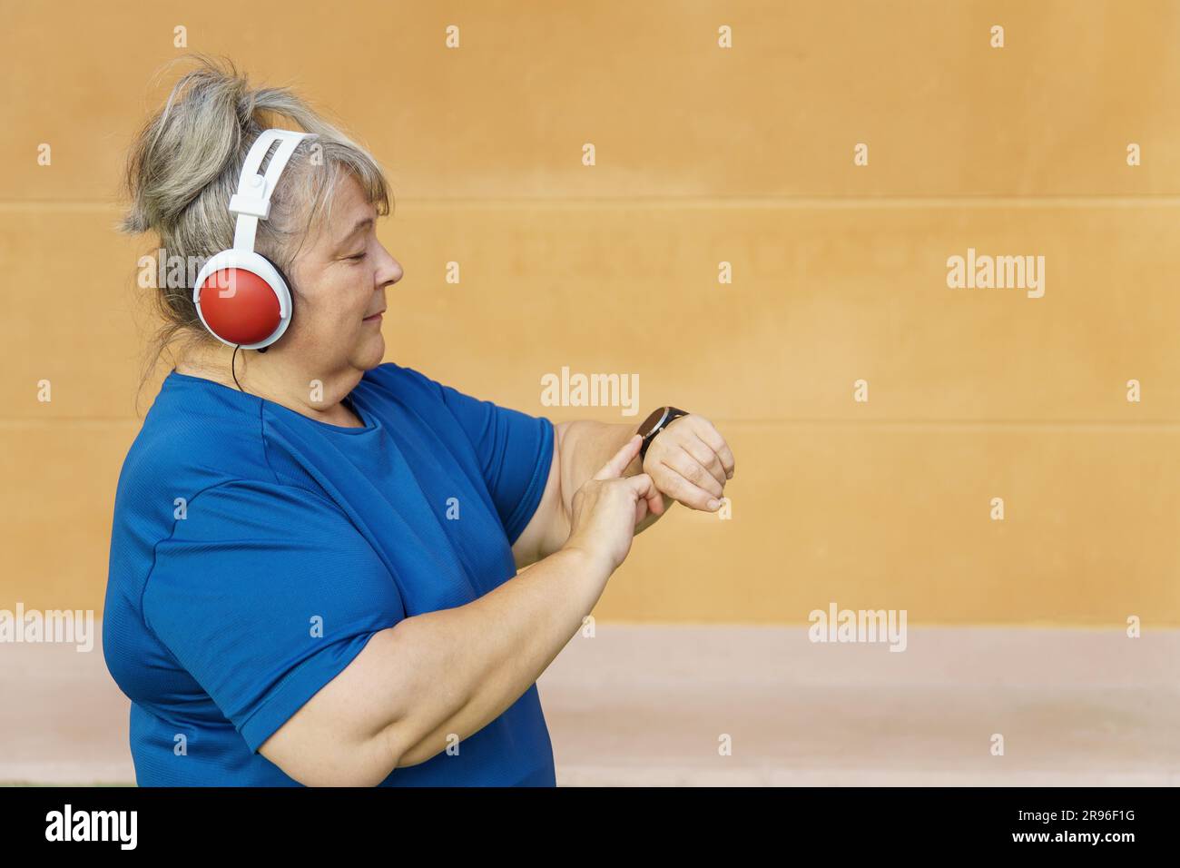 Una donna più anziana, con abbigliamento sportivo e cuffie in sovrappeso, si rivolge al suo smartwatch mentre si allena nel parco Foto Stock