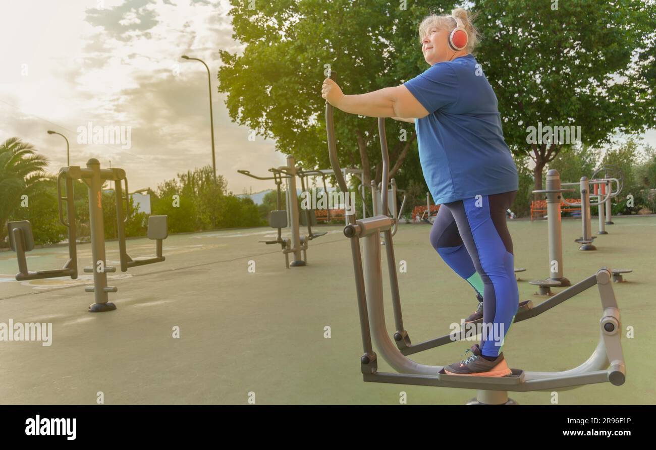 Donna anziana obesa attiva in abbigliamento sportivo che si allena su una macchina da parcheggio mentre ascolta la musica sulle cuffie Foto Stock