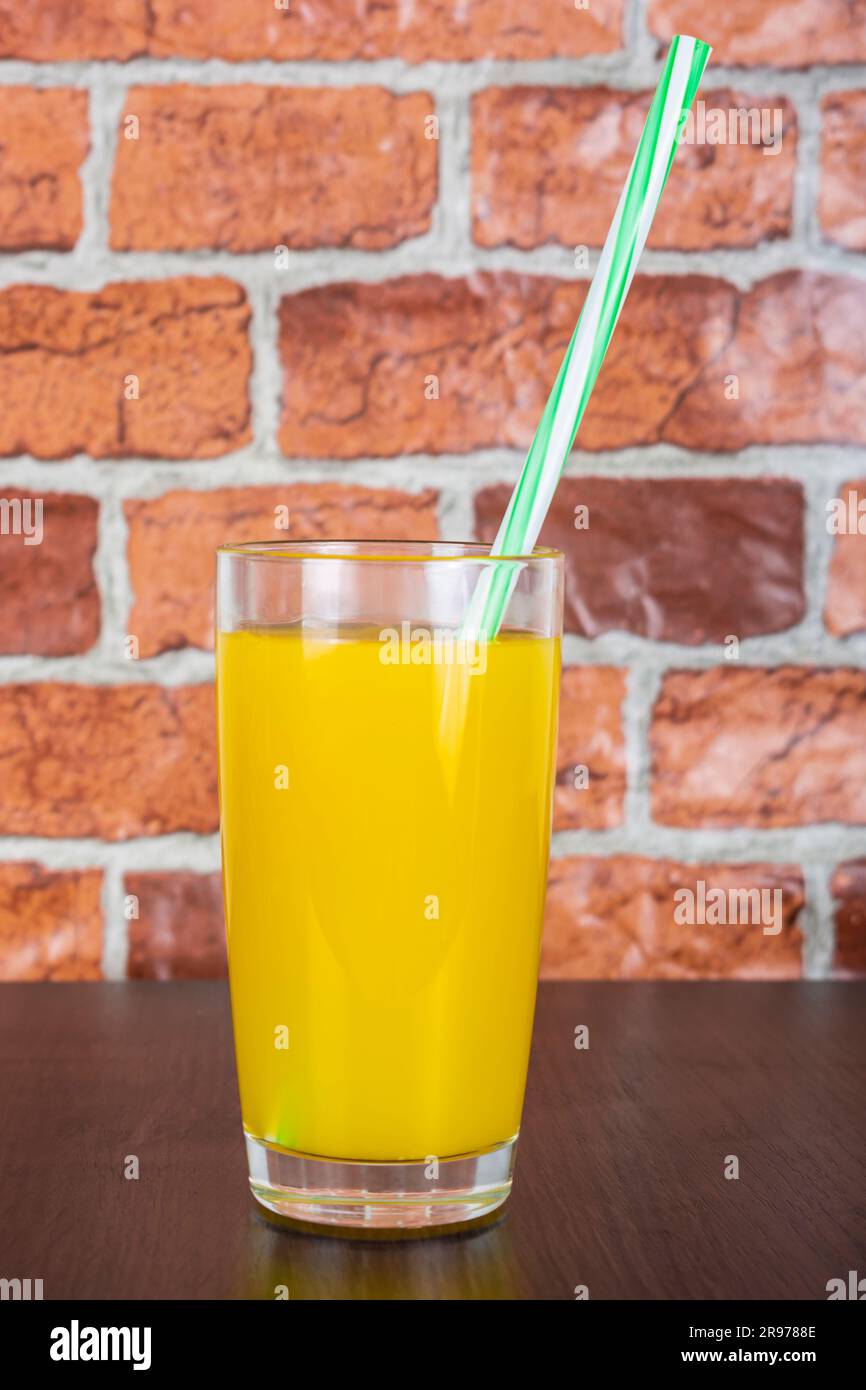 Succo d'arancia in una tazza di vetro con un tubo su un tavolo di legno e uno sfondo di una parete di mattoni arancioni Foto Stock