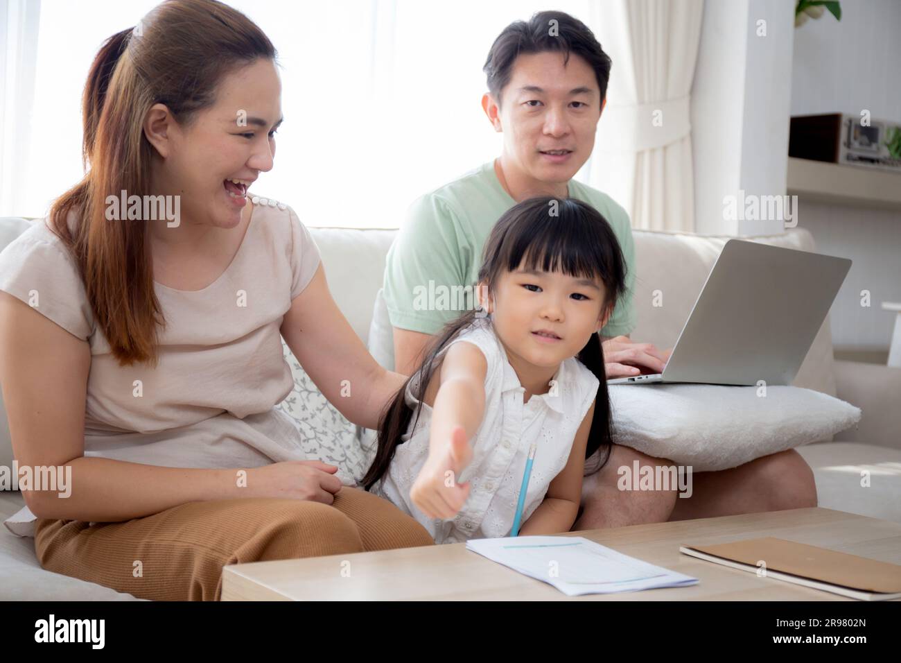 Famiglia felice con la madre che insegna i compiti a casa con la figlia e il padre che lavora con il portatile sul divano nel soggiorno di casa, mamma che spiega il lavoro scolastico con Foto Stock