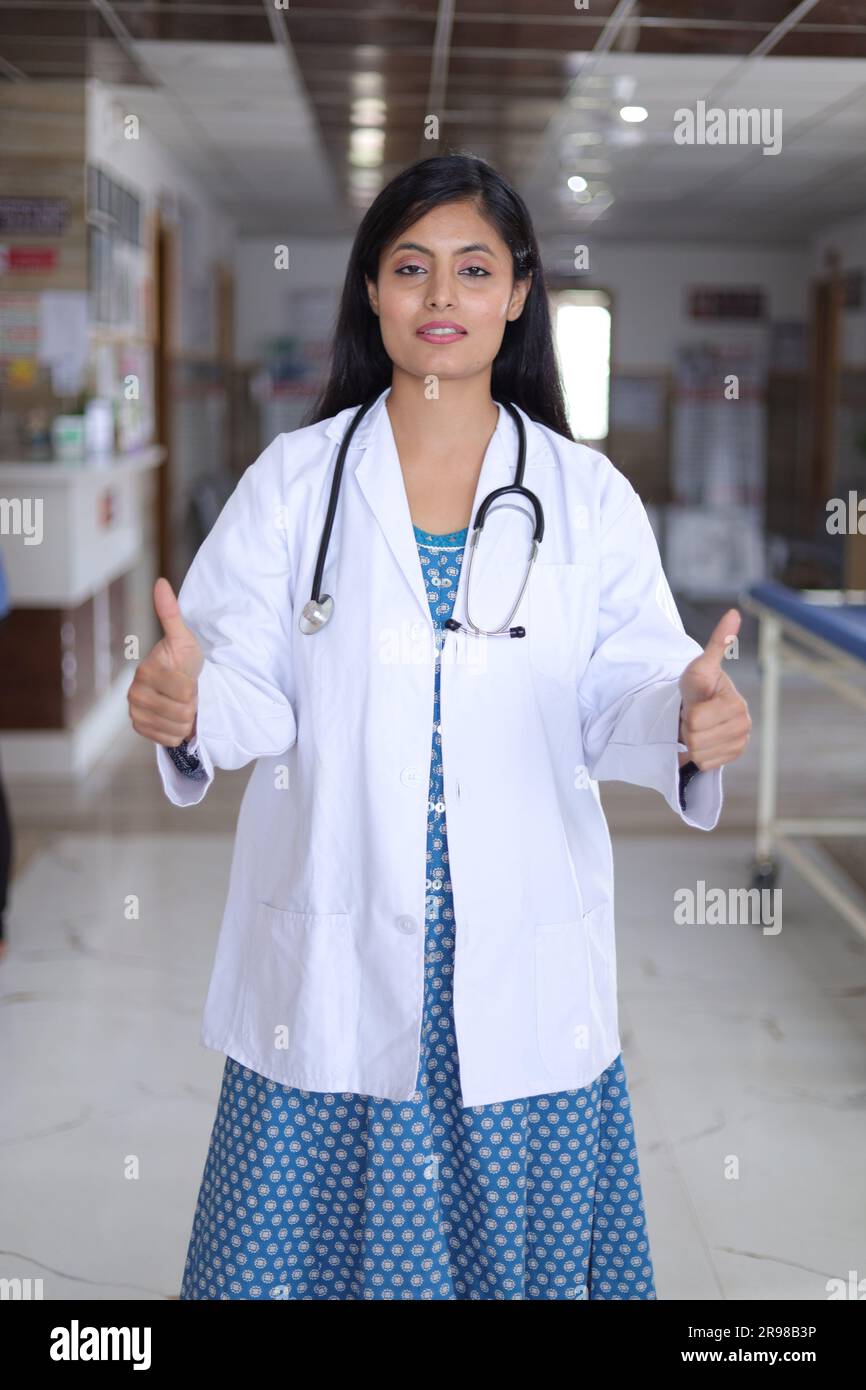 Ritratto di una dottoressa che mostra il pollice in alto. Infermiera felice in uniforme blu in piedi nel corridoio dell'ospedale sullo sfondo. Foto Stock