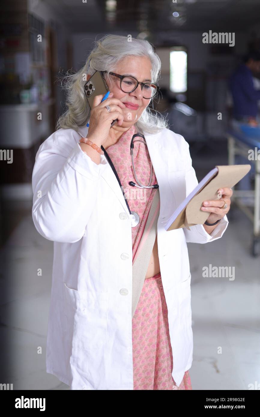 Ritratto di una donna anziana intelligente in camice bianco da laboratorio che parla durante una telefonata con un blocco note in mano. blocco di scrittura. Foto Stock