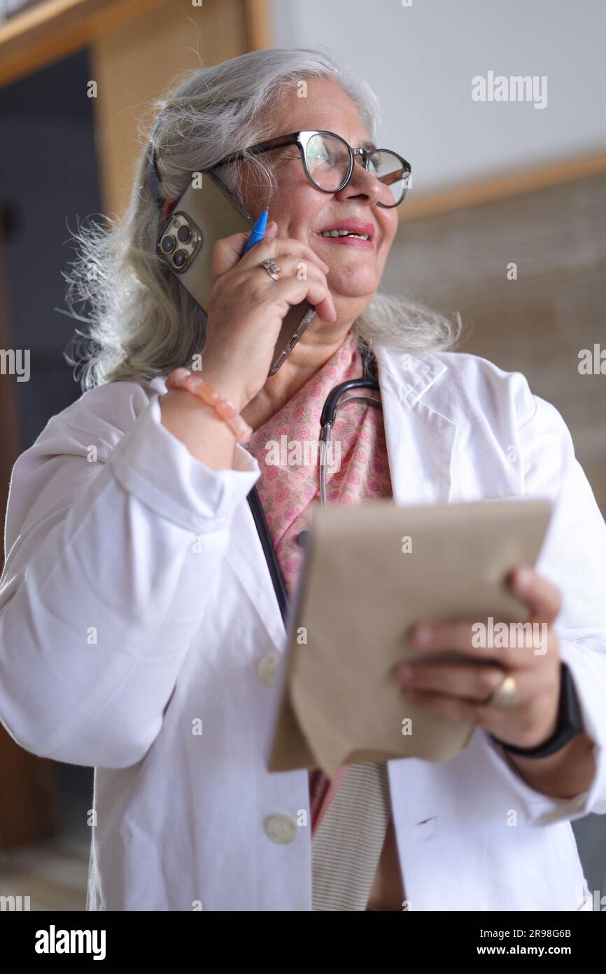 Ritratto di una donna anziana intelligente in camice bianco da laboratorio che parla durante una telefonata con un blocco note in mano. blocco di scrittura. Foto Stock