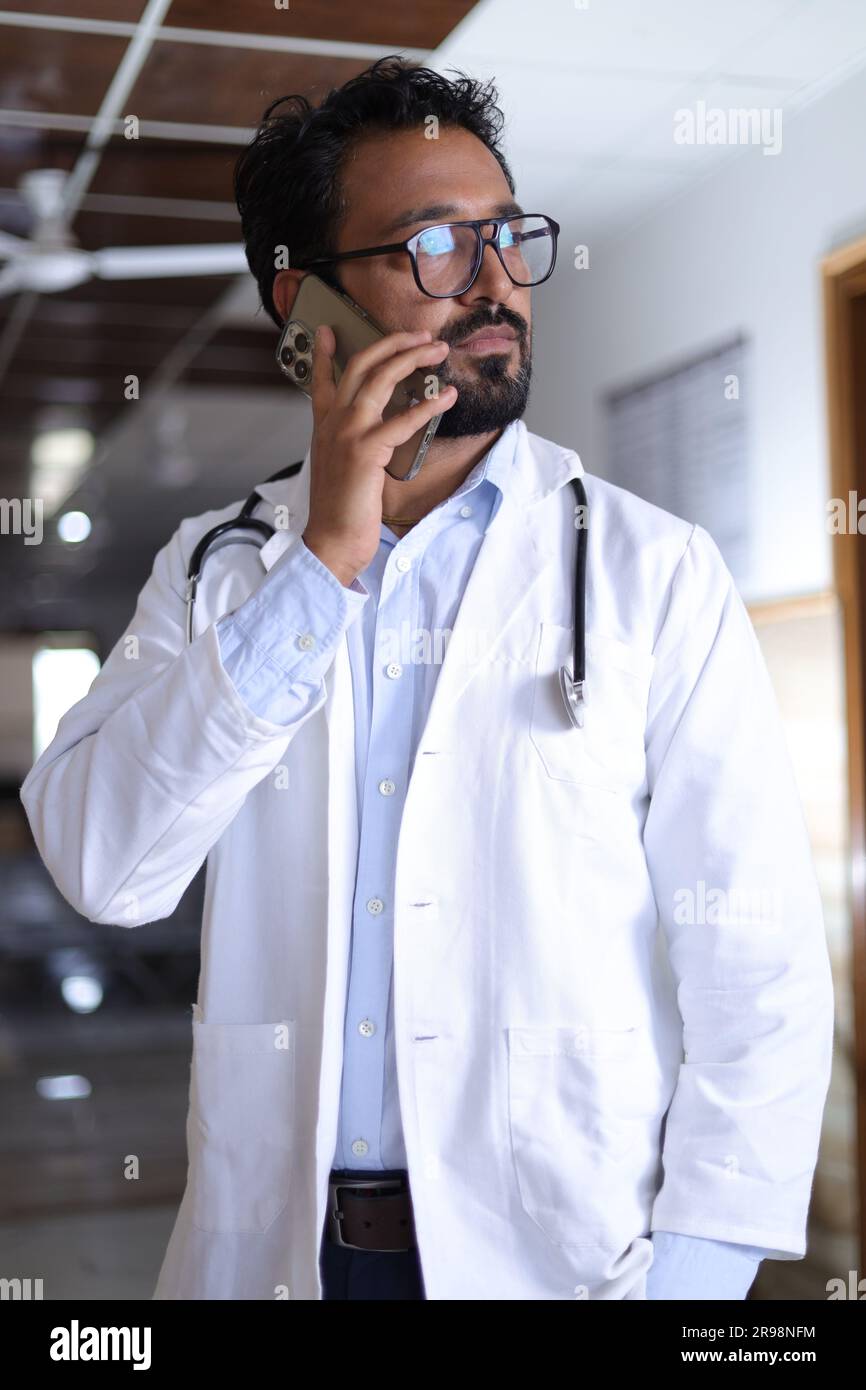 Ritratto medico professionale in corridoio di ospedale vestito in scrub e camici da laboratorio, con stetoscopi. Indiano decente che parla su un cellulare. Foto Stock