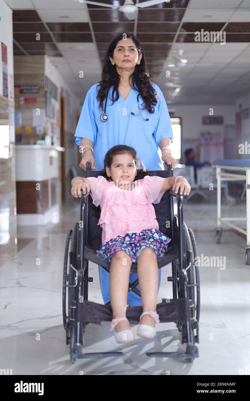 bambina piccola seduta su una sedia a rotelle con un'infermiera che si prende cura di lei, entrambi sono in piedi nel corridoio dell'ospedale, lesioni, disabilità fisica. Foto Stock