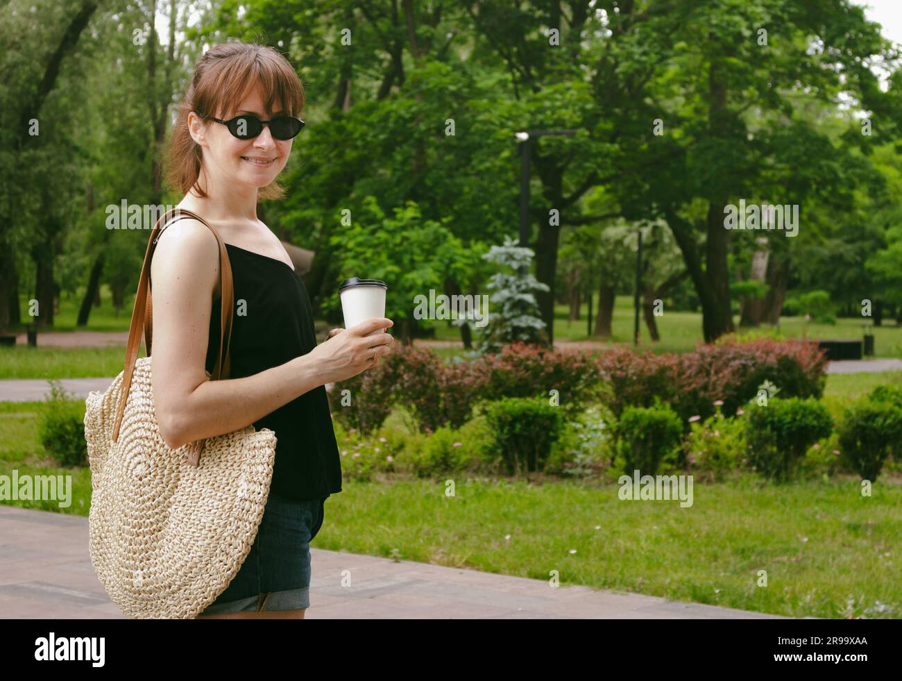 Ragazza sorridente con una tazza di caffè nel parco. Bella donna con gli occhiali da sole che cammina nel parco. Informazioni generali sull'ora legale. Weekend estivo. Felice freelance. Foto Stock