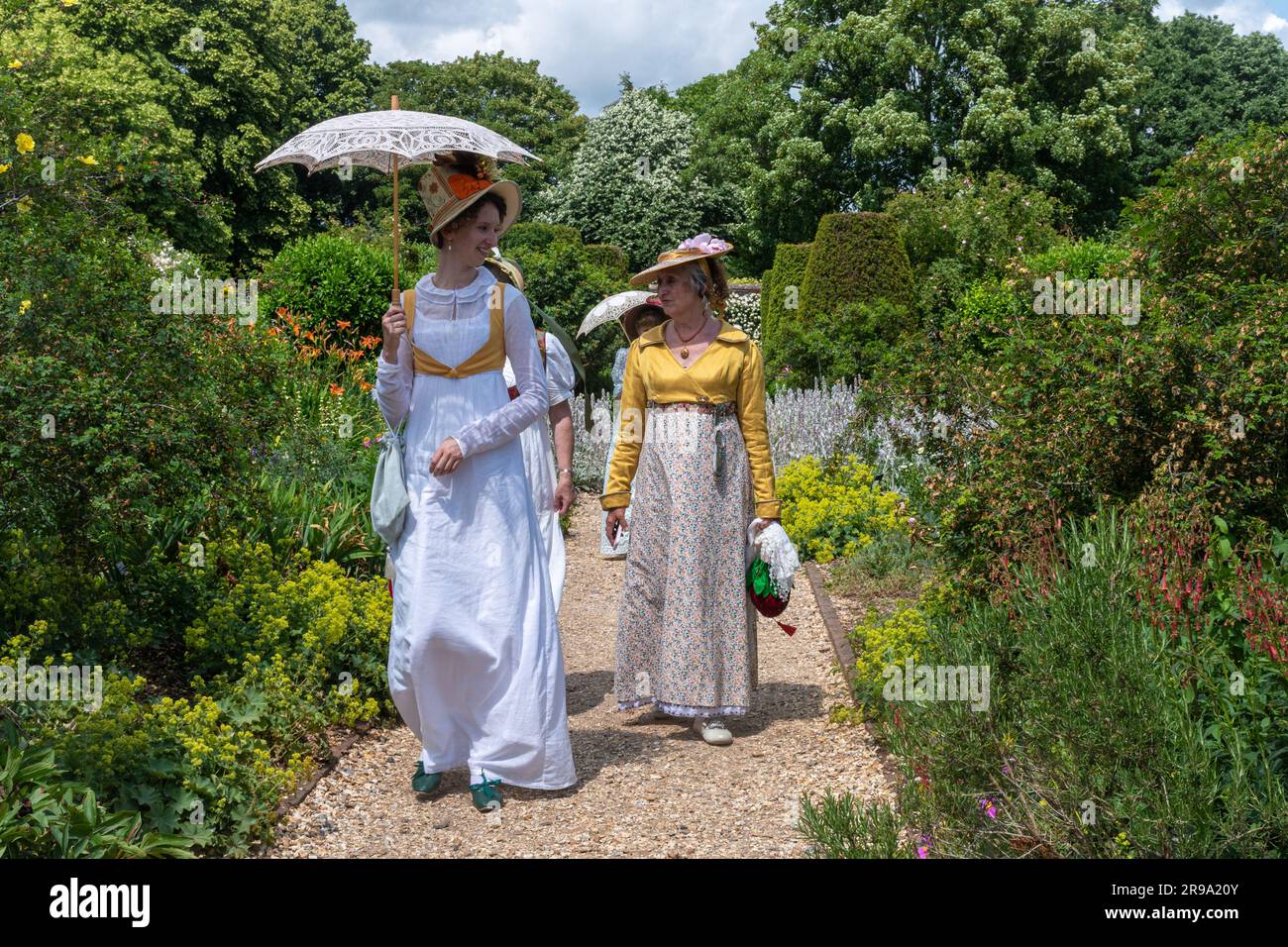 Signore vestite con abiti o costumi vittoriani d'epoca che camminano attraverso un giardino inglese in estate trasportando ombrelloni, Inghilterra, Regno Unito Foto Stock