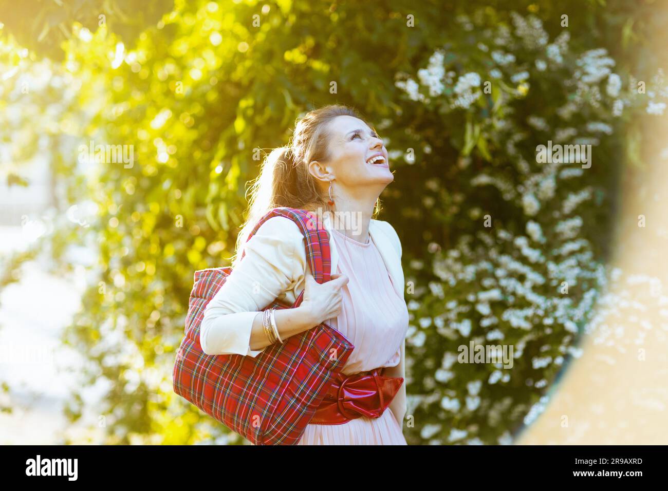 felice elegante donna di 40 anni in abito rosa e giacca bianca in città con borsa rossa. Foto Stock