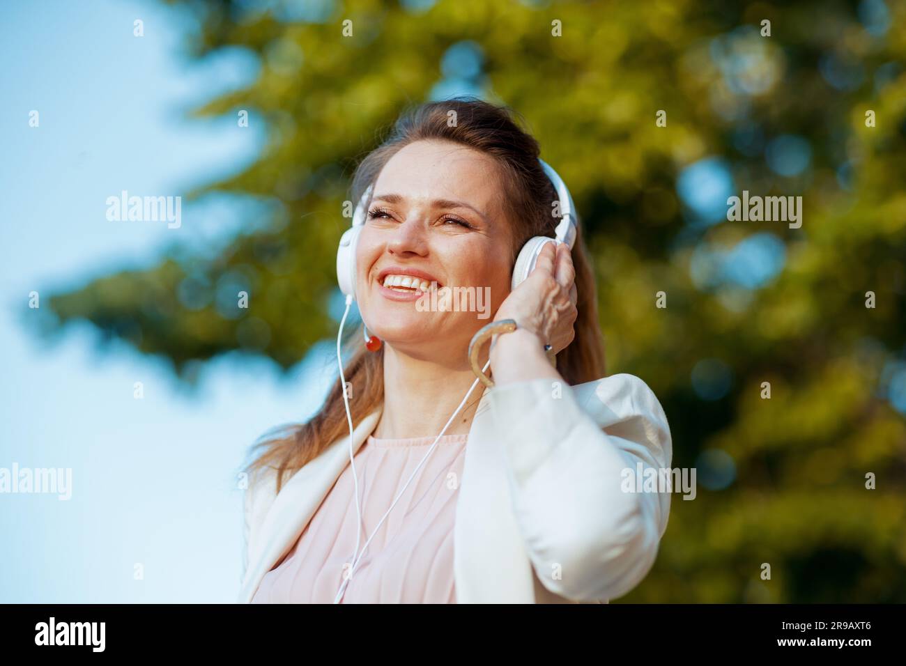 elegante donna sorridente di 40 anni con abito rosa e giacca bianca in città che ascolta la musica con le cuffie. Foto Stock