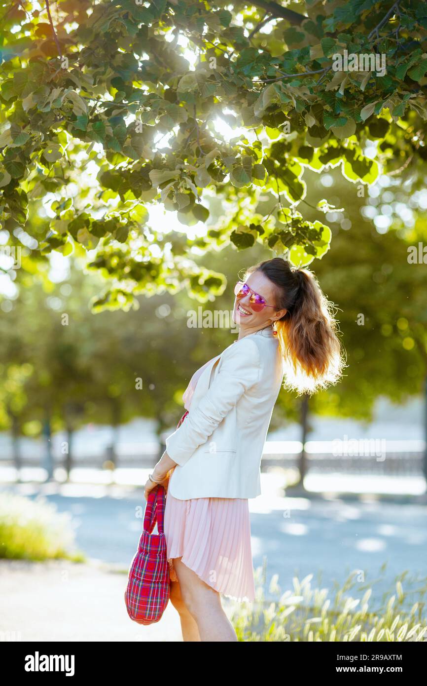 felice ed elegante donna di 40 anni in abito rosa e giacca bianca in città con borsa rossa e occhiali da sole. Foto Stock