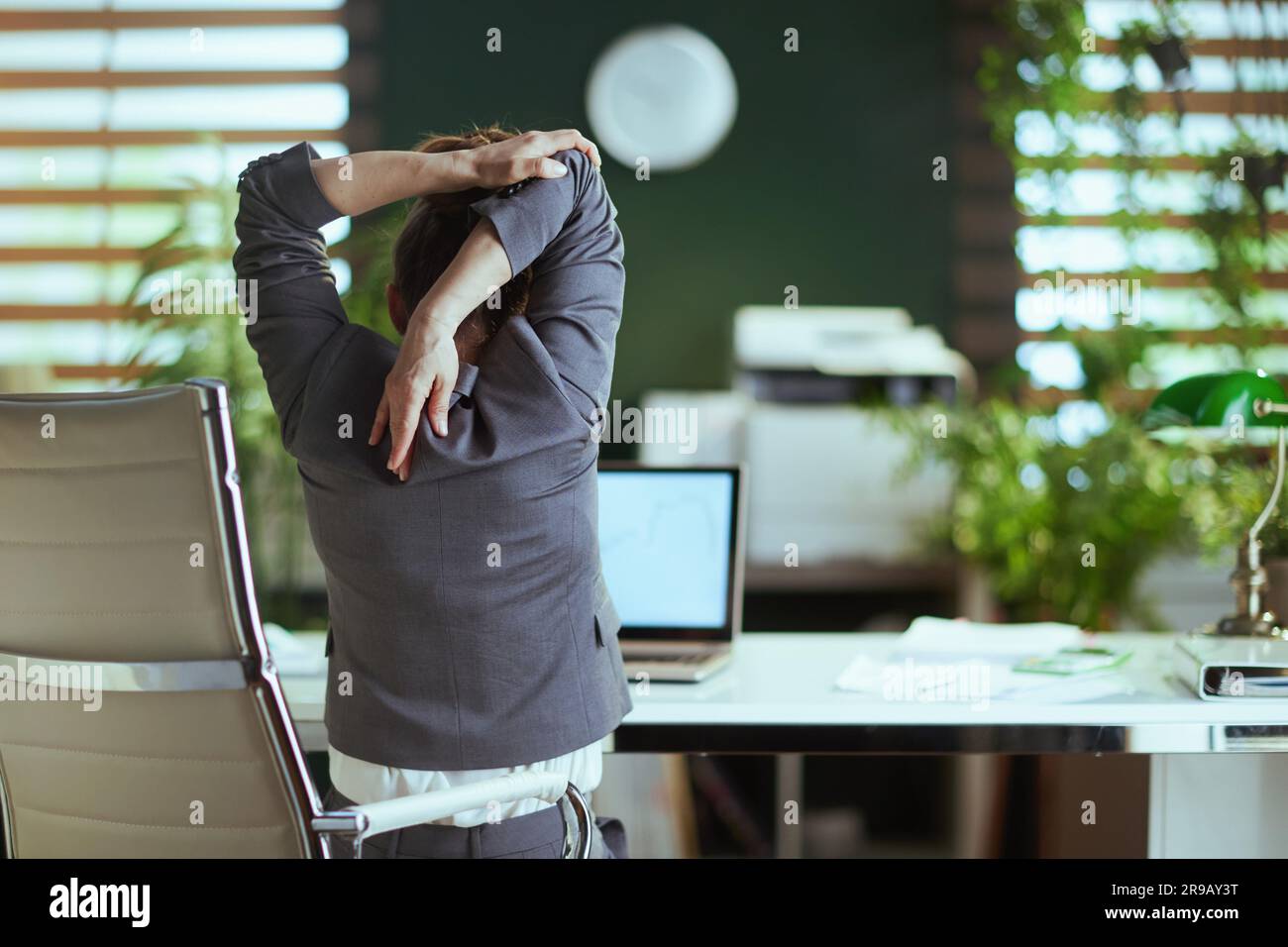 Ambiente di lavoro sostenibile. Visto da dietro moderna donna di affari in un vestito grigio di affari in moderno verde ufficio che stretching mano. Foto Stock
