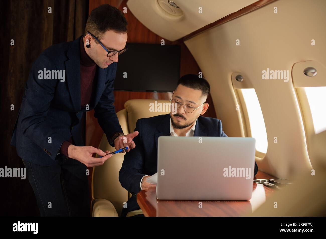 Elegante coppia di uomini d'affari seduti all'interno di un jet privato e che parla di lavoro mentre si è in volo guardando un laptop, un viaggio e una compagnia aerea Foto Stock