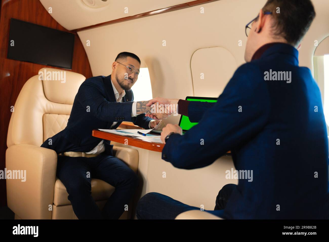 Due coppie di uomini d'affari sedute all'interno di un aereo privato a reazione e parlando di lavoro che stringono la mano, mentre si trova in volo il concetto di business della compagnia aerea Travel Foto Stock