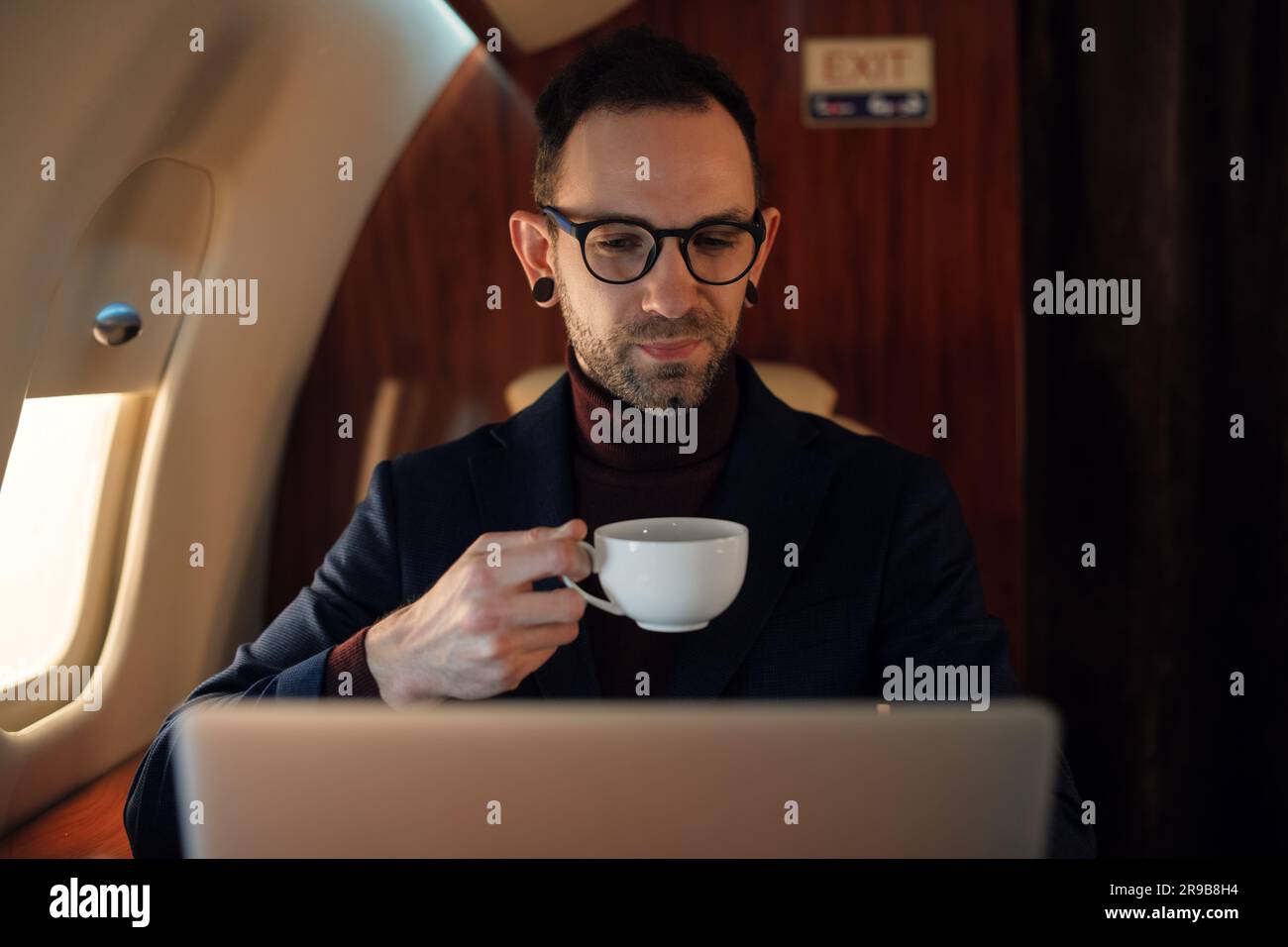 Elegante giovane CEO uomo d'affari con occhiali da vista seduto all'interno di un aereo privato che si gode un caffè mentre lavora sul suo moderno computer portatile Foto Stock