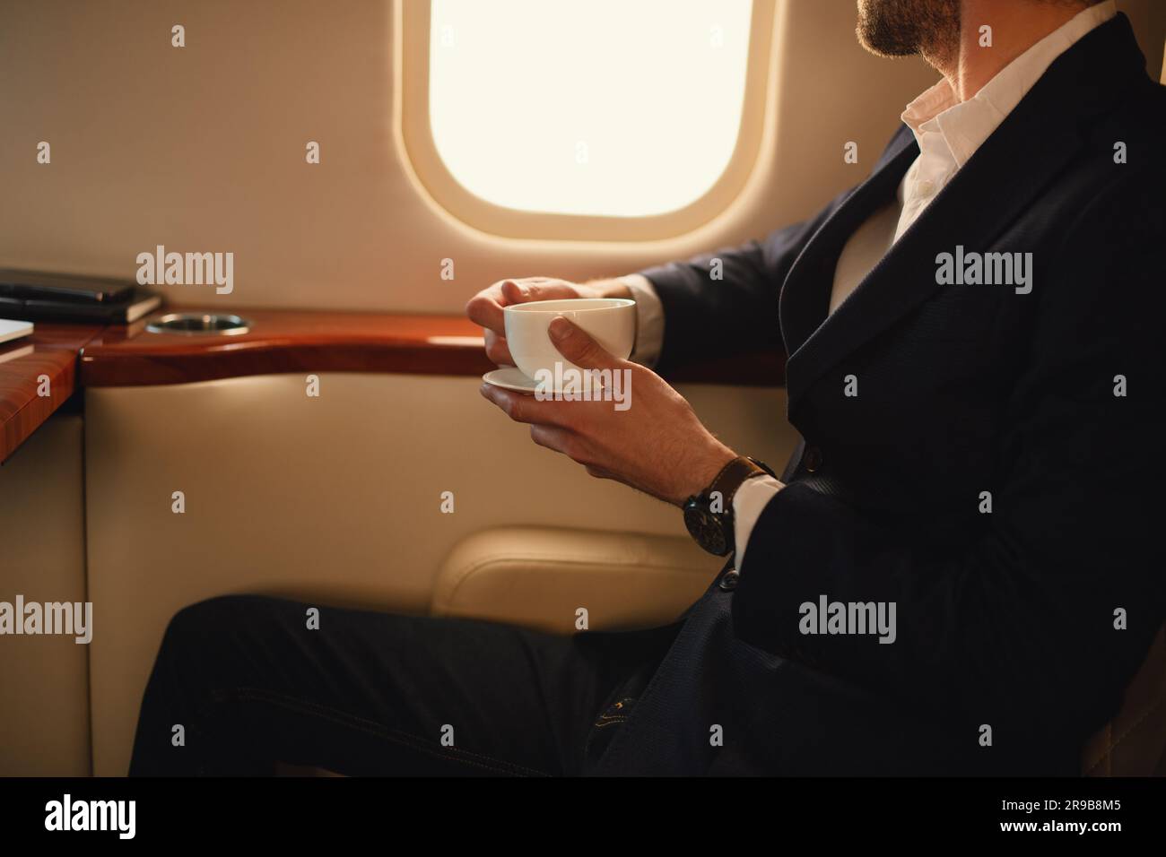 Irriconoscibile elegante uomo d'affari CEO con occhiali da vista in una giacca blu seduto all'interno di un jet privato di un aereo che beve un caffè guardando da una finestra Foto Stock