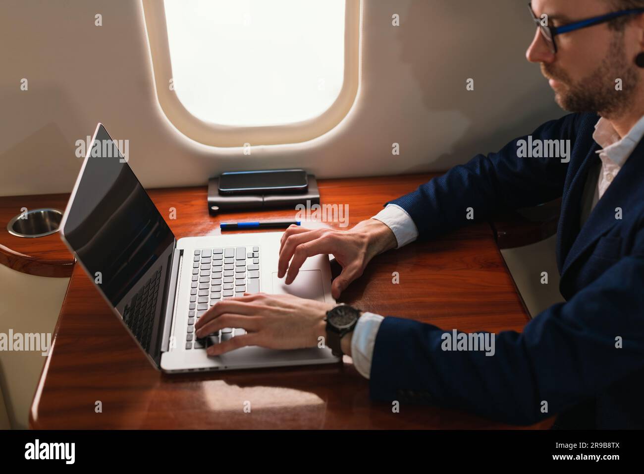 Giovane ed elegante uomo d'affari CEO con occhiali da vista in una giacca blu che utilizza un moderno laptop da lavoro in un jet privato di prima classe con vista laterale Foto Stock