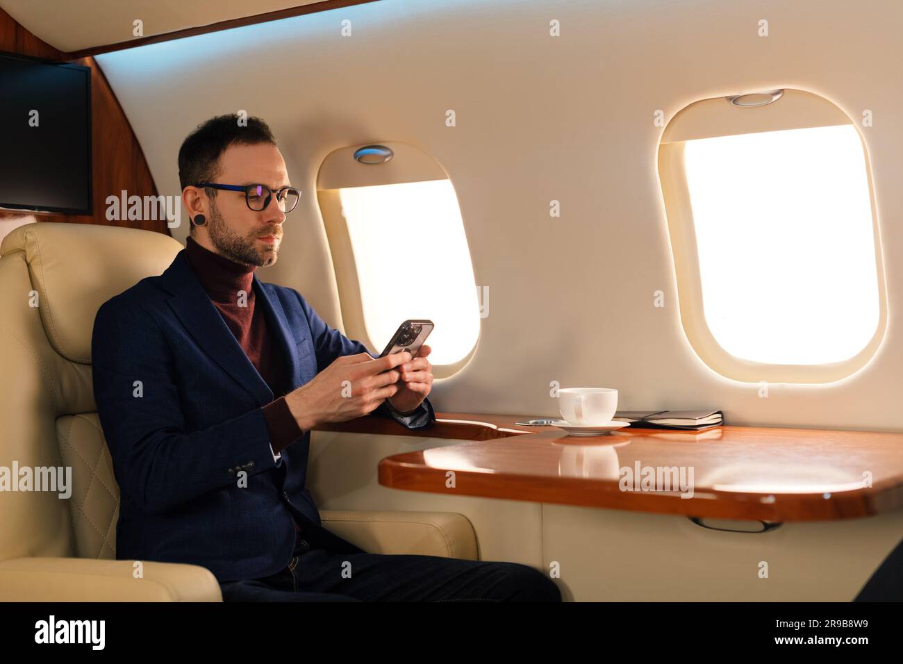 Elegante giovane CEO giovane uomo d'affari con occhiali da vista seduto all'interno in un jet privato di prima classe mentre si lavora con uno smartphone moderno Foto Stock