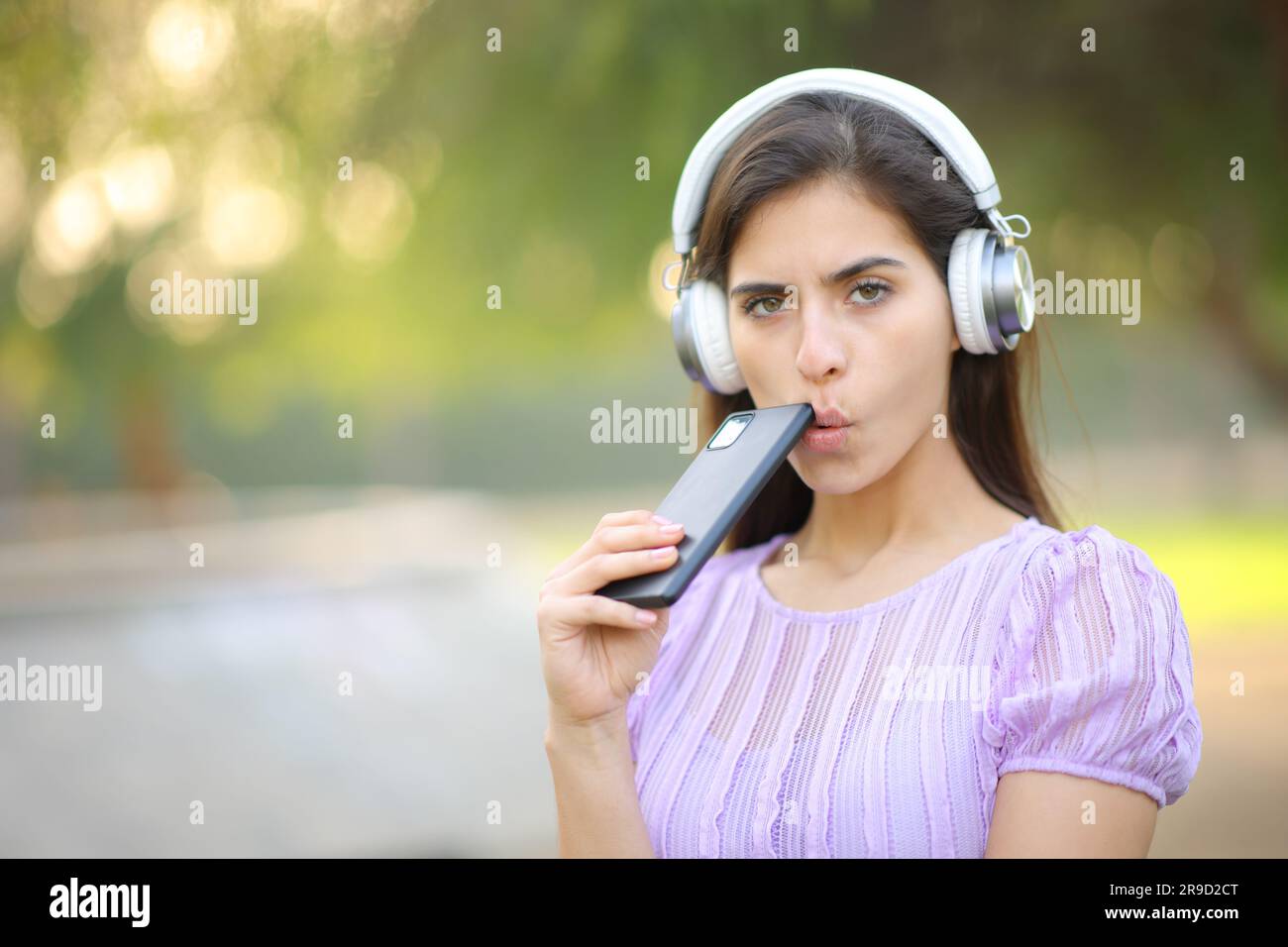 Donna sospetta che ti guarda mentre ascolti audio digitale con cuffie e smartphone in un parco Foto Stock