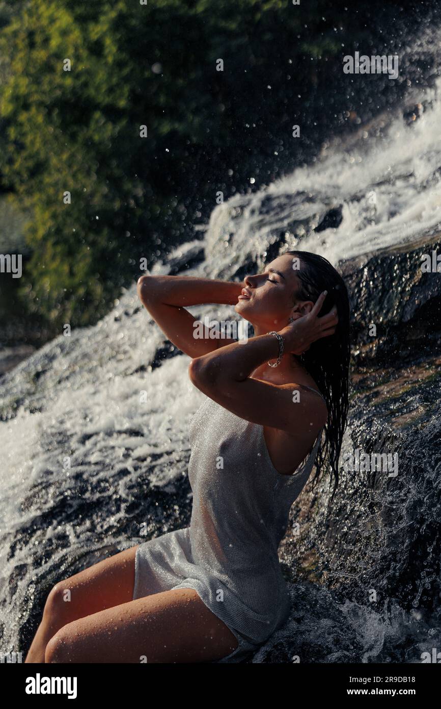 Giovane donna in abito bagnato seduta sulla roccia e con gli occhi chiusi vicino alla cascata tra flussi d'acqua, spruzzi e schiuma. Primo piano. Foto Stock