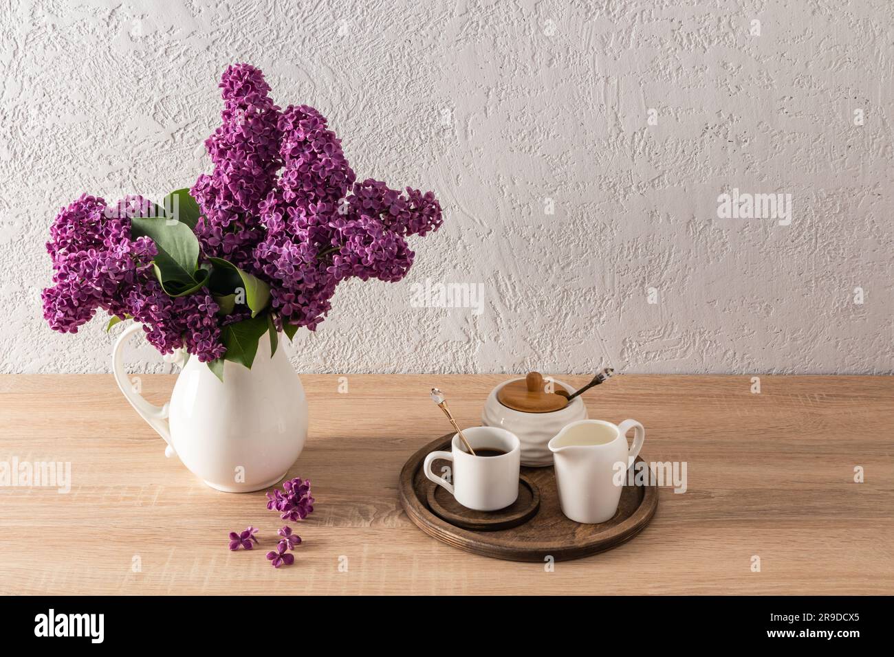 Primavera natura morta. Una caraffa con un bouquet di lilaci e un vassoio rotondo in legno con una tazza di caffè mattutino, una ciotola da zucchero su un ripiano in legno Foto Stock