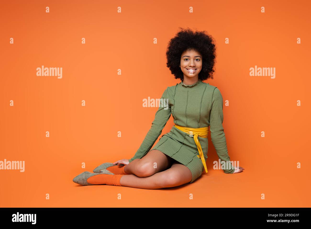 Allegra e adolescente ragazza afroamericana in elegante abito verde sorridente davanti alla macchina fotografica mentre si siede e posa su sfondo arancione, cultura giovanile e.. Foto Stock