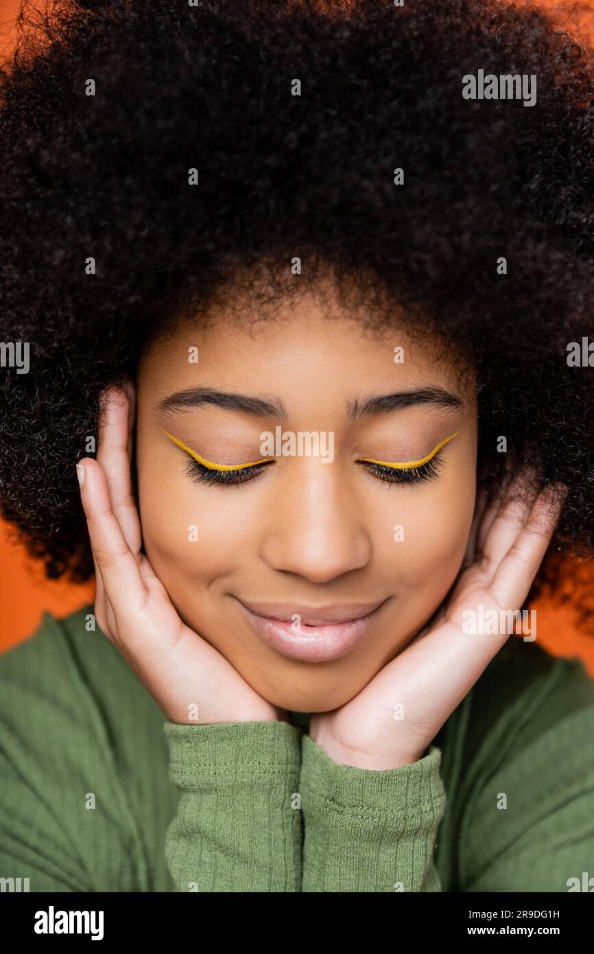 Ritratto di una ragazza afro-americana adolescente sorridente e alla moda con un trucco luminoso che tocca le guance e posa isolata sull'arancio, la cultura giovanile e i generi Foto Stock
