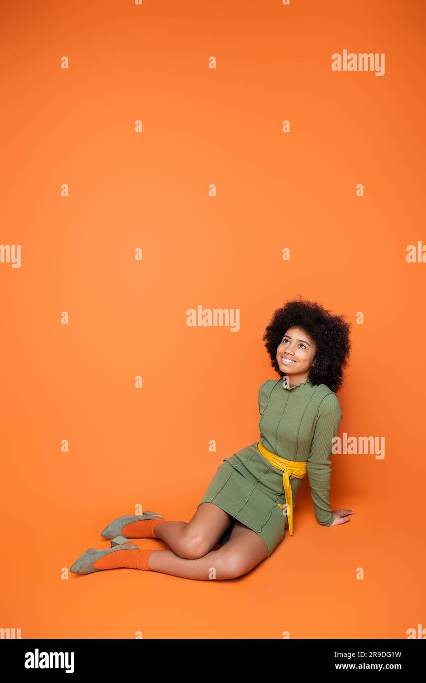 Un'intera lunghezza di adolescenti afro-americani allegri e alla moda in abito verde che guarda in alto mentre si siede e posa su sfondo arancione, cultura giovanile e Foto Stock