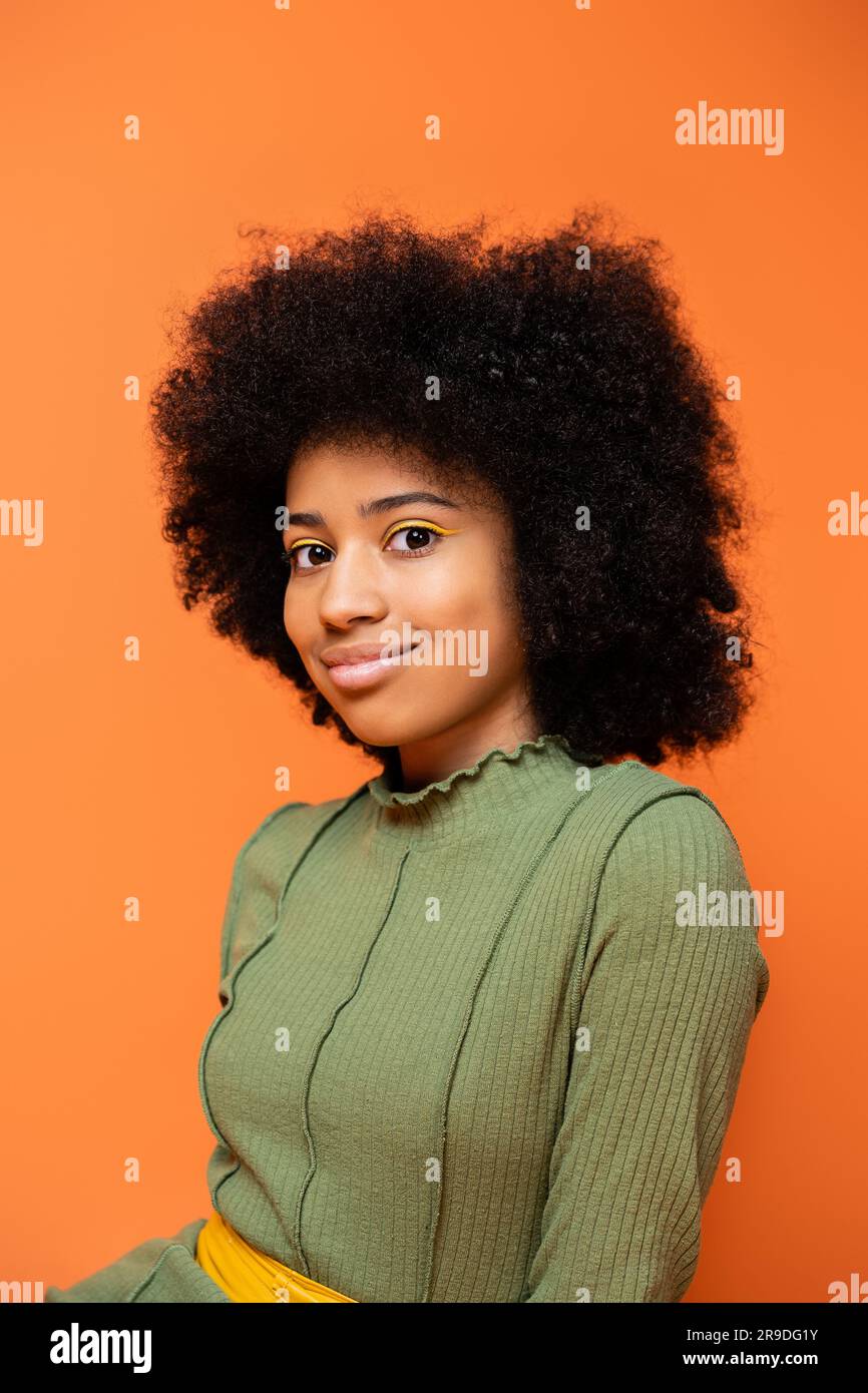 Ritratto di una ragazza afro-americana alla moda con un audace trucco che indossa un vestito verde e sorride alla macchina fotografica isolata su arancione, cultura giovanile e ge Foto Stock