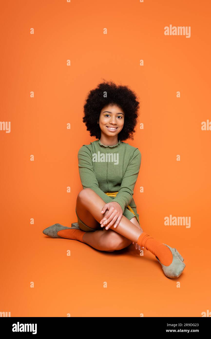 Un modello afro-americano adolescente con un trucco colorato che indossa un vestito verde e che siede su sfondo arancione, cultura giovanile e gene Foto Stock