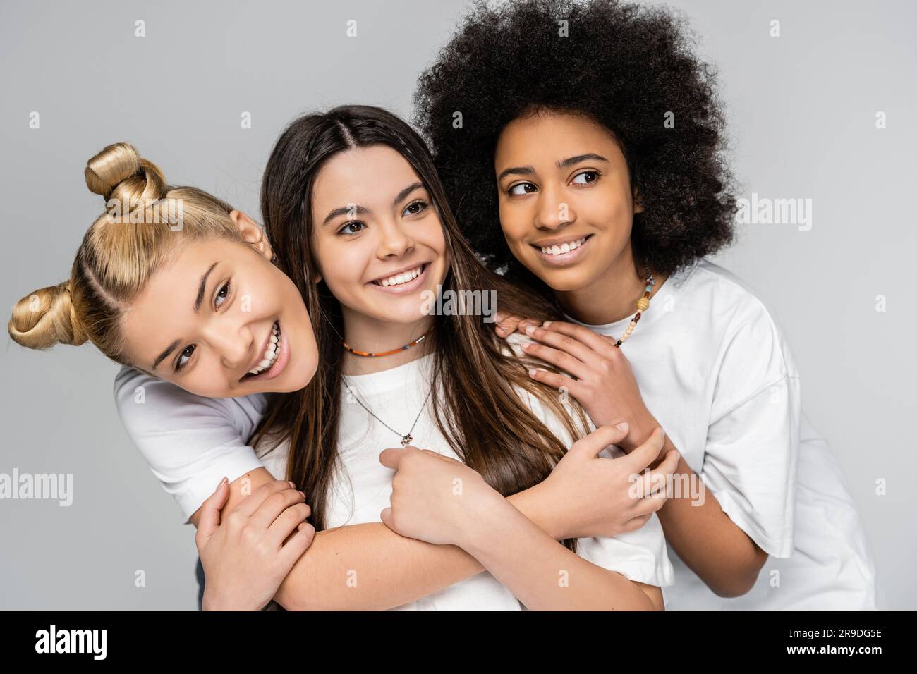 Ritratto di adolescenti allegri e multietnici in t-shirt bianche che abbracciano e guardano lontano mentre si posa isolata su ragazze grigie, adolescenti e generat Foto Stock