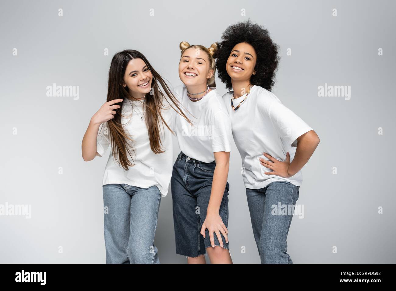 Ragazze adolescenti eleganti e gioiose in jeans e t-shirt bianche che guardano la fotocamera mentre posano insieme su sfondo grigio, modelli per adolescenza e. Foto Stock