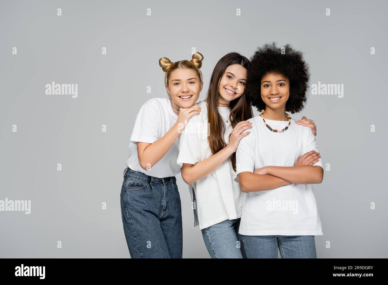 Ragazze positive e adolescenti con t-shirt e jeans bianchi che abbracciano una fidanzata afroamericana sicura di sé che incrocia le braccia mentre si trova isolata sul grigio, li Foto Stock