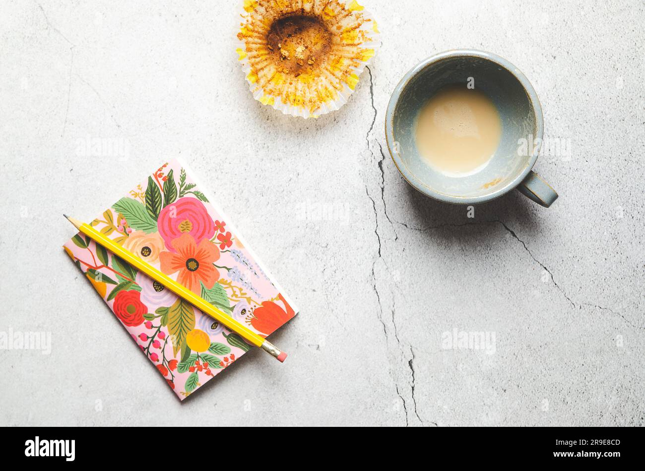 Un notebook fiorito con una matita gialla, una tazza vuota di latte e una tazza da forno, su sfondo grigio. Foto Stock