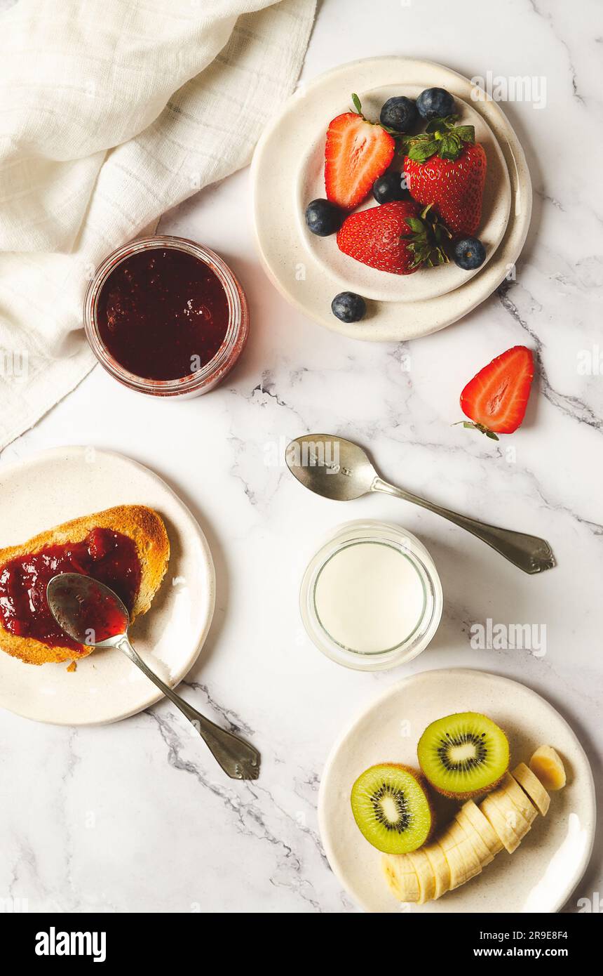 Yogurt semplice, frutta fresca, marmellata di fragole in un vaso e su una fetta di pane tostato con un tovagliolo bianco su fondo marmorizzato. Vista dall'alto. Foto Stock
