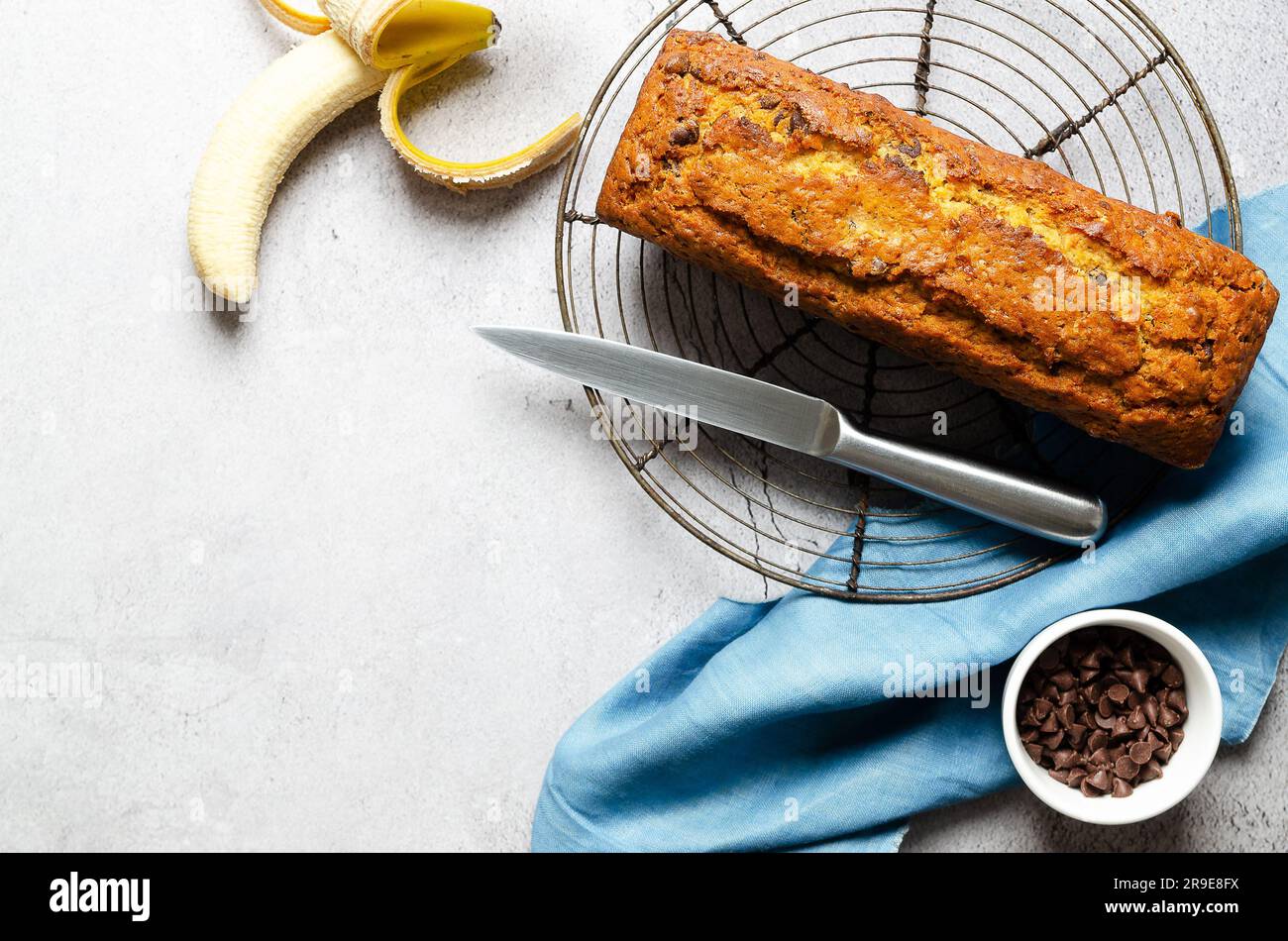 Un pane alla banana con gocce di cioccolato, una banana, gocce di cioccolato in una ciotola e un tovagliolo blu, su sfondo grigio. Foto Stock