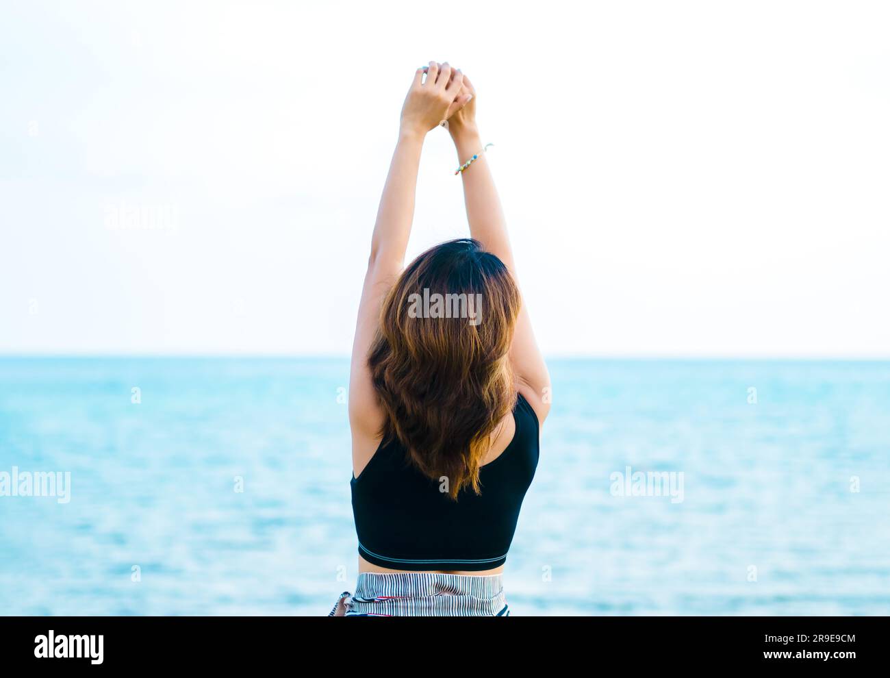 Schiena di donna asiatica in canotta nera con occhi di chiusura con rilassante alzare le mani e distendersi sullo sfondo della spiaggia. Felice femmina in piedi di fronte a te Foto Stock