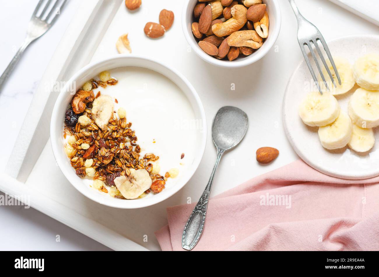 Yogurt con muesli, banana, noci e uva passa e un tovagliolo rosa su un vassoio bianco Foto Stock