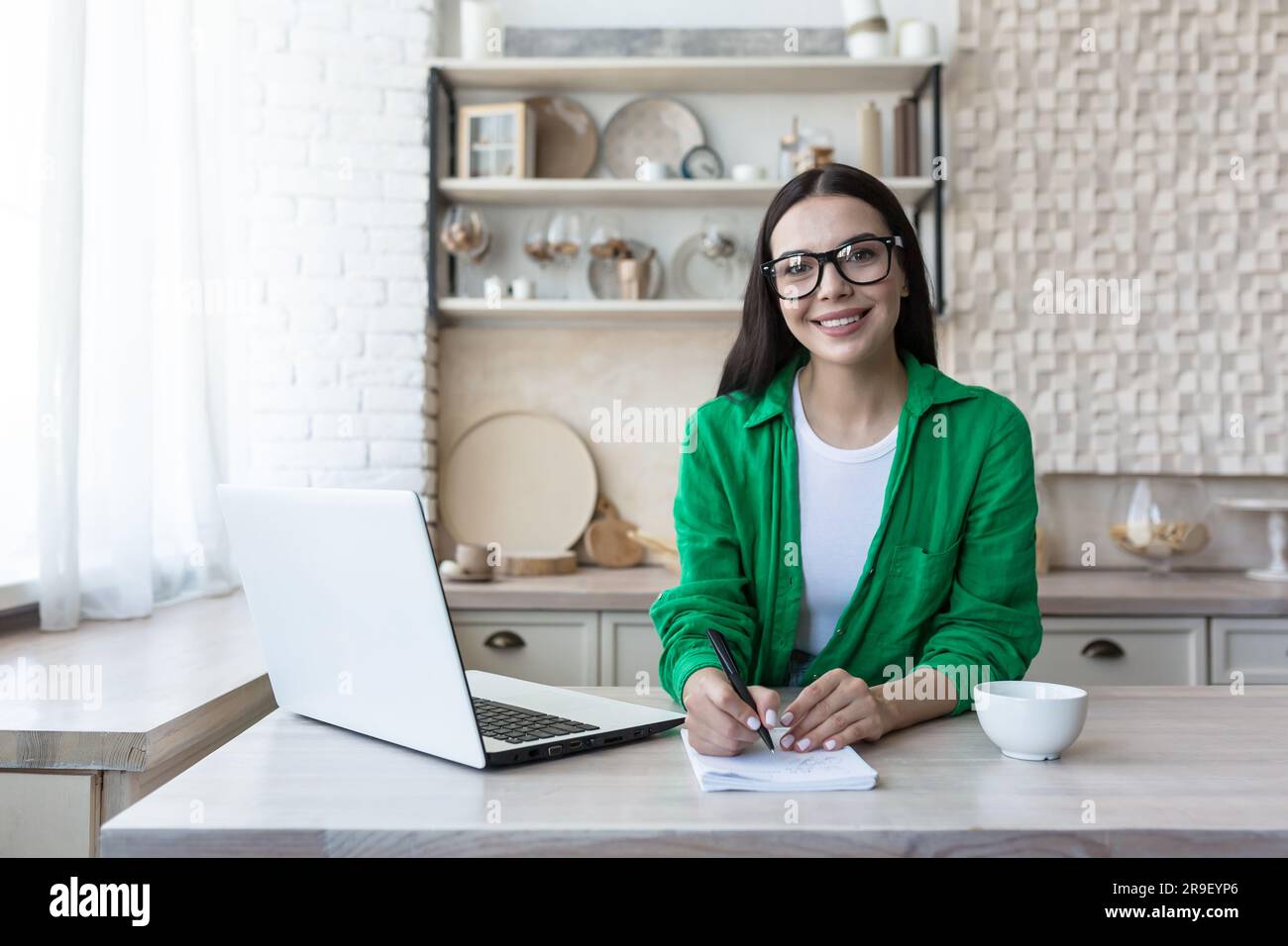 Ritratto di una giovane studentessa che studia in remoto con un notebook. Seduto in cucina al tavolo con un notebook e guardando sorridente la fotocamera. Foto Stock