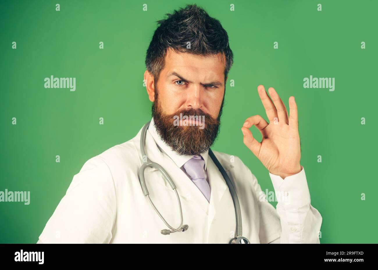 Un medico serio che mostra un segno positivo. Uomo barbuto con stetoscopio in camice medico bianco con approvazione di firma. Medicina, clinica, trattamento, assistenza sanitaria Foto Stock