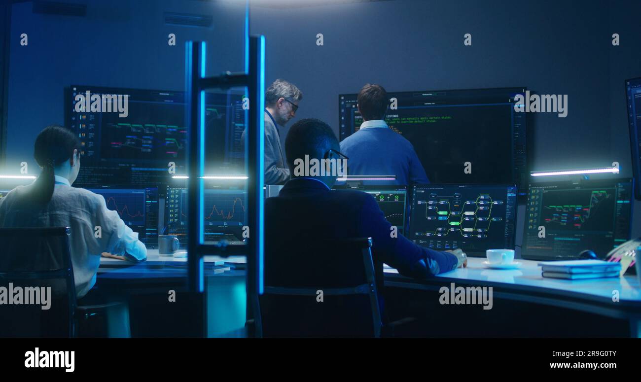 Team multietnico di specialisti tecnici IT e programmatori software lavora con server dati, grafici di analisi e rete blockchain su computer e grandi schermi digitali in ufficio di monitoraggio di notte. Foto Stock